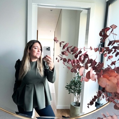 Blusa verde oliva com plissados e mangas volumosas, vista ao espelho num ambiente interior com planta e decoração