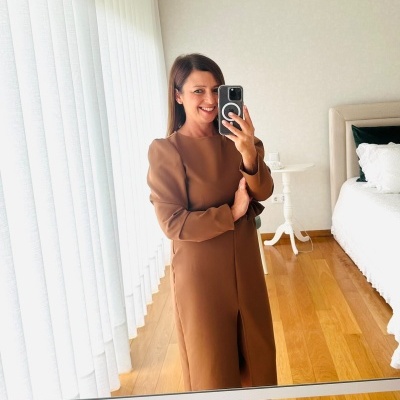 Mulher com vestido castanho comprido e sapatos rosa claro a tirar selfie em quarto com chão de madeira e cortinas brancas.