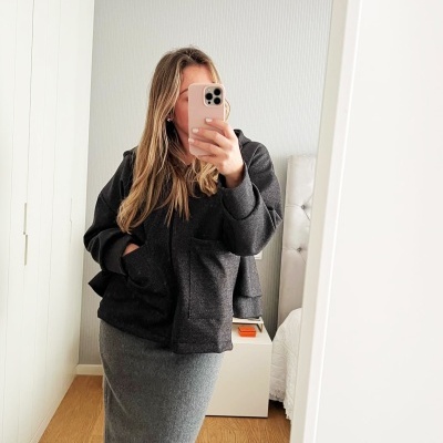 Mulher a tirar selfie usando casaco preto e saia comprida cinza em quarto com chão de madeira