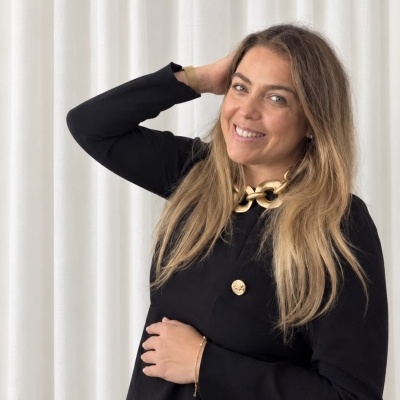 Mulher com blazer preto, colar e pulseira dourados em fundo branco.