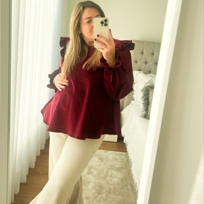 Mulher a tirar selfie usando blusa bordô e calças creme num quarto