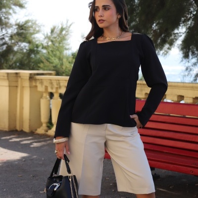 Mulher com blusa preta, bermuda bege e bolsa preta ao ar livre junto a banco vermelho