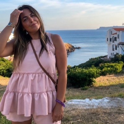 Mulher com vestido rosa claro com babados, pulseira roxa e bracelete prateado junto ao mar