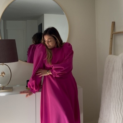 Mulher com vestido rosa fúcsia de mangas bufantes perto de móvel branco com espelho redondo ao fundo