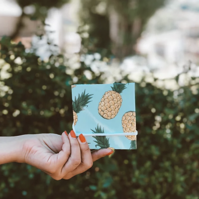 Carteira azul clara com padrão de abacaxis amarelos e folhas verdes sendo segurada por mãos com unhas laranja