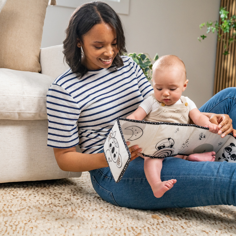 Mulher com bebé a olhar um livro de pano preto e branco sentado no chão sobre tapete claro
