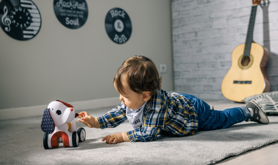 Criança a brincar com cão de brinquedo vermelho, branco e azul num tapete cinzento com discos de vinil e guitarra ao fundo