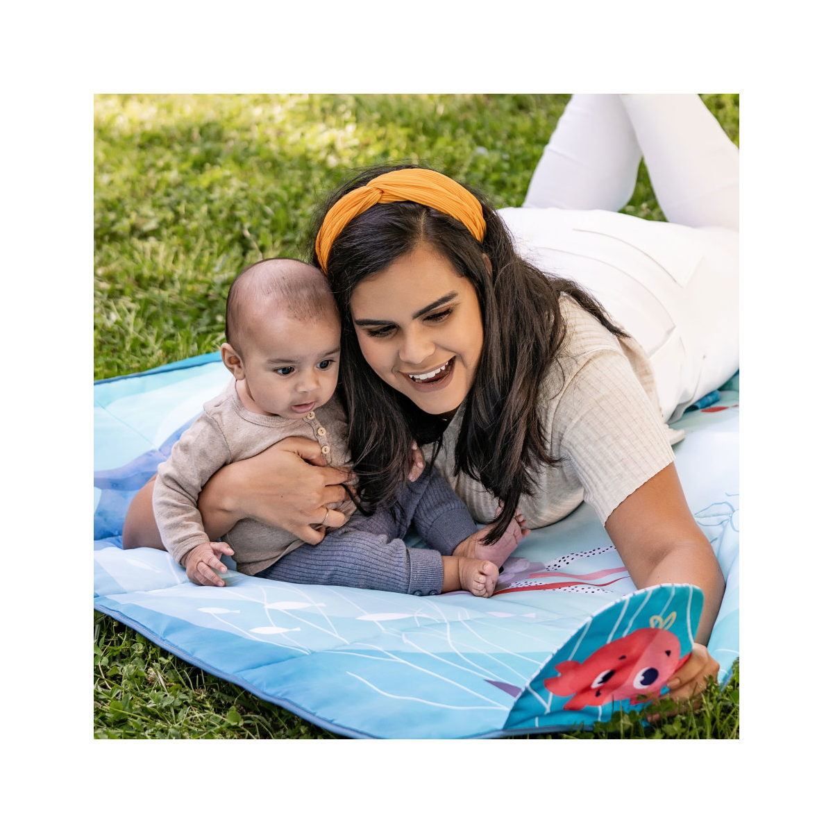 Bebé e mulher numa manta azul com desenho de caranguejo vermelho no relvado