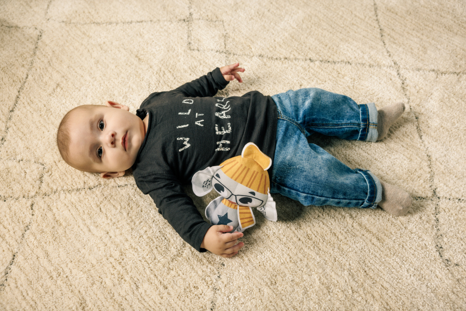 Bebé deitado num tapete bege com camisola preta e calças de ganga azuis a segurar um brinquedo de tecido