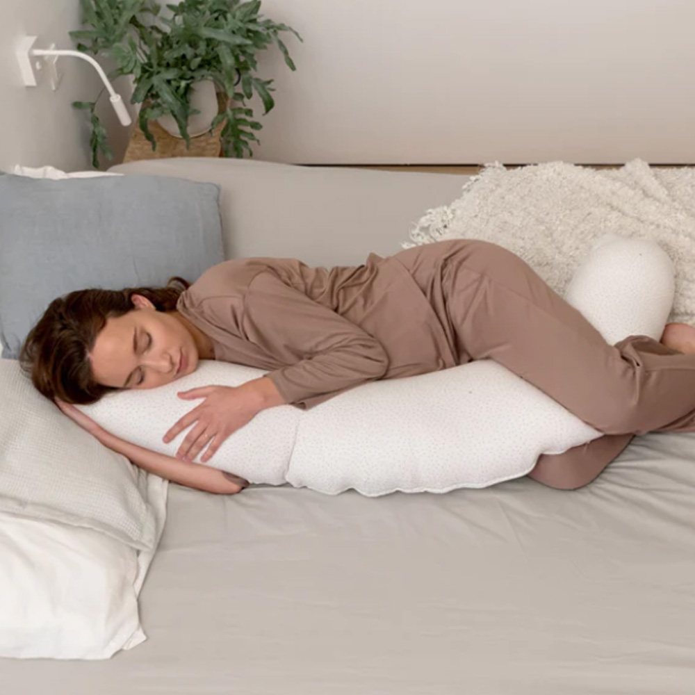 Mulher de pijama castanho claro deitada e abraçando travesseiro branco de corpo largo sobre cama cinza