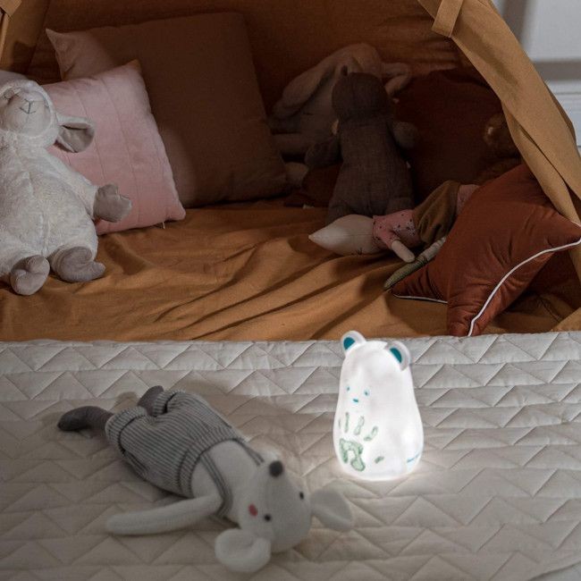 Quarto infantil com tenda castanha, almofadas, brinquedos de pelúcia e urso luminoso sobre colcha branca acolchoada
