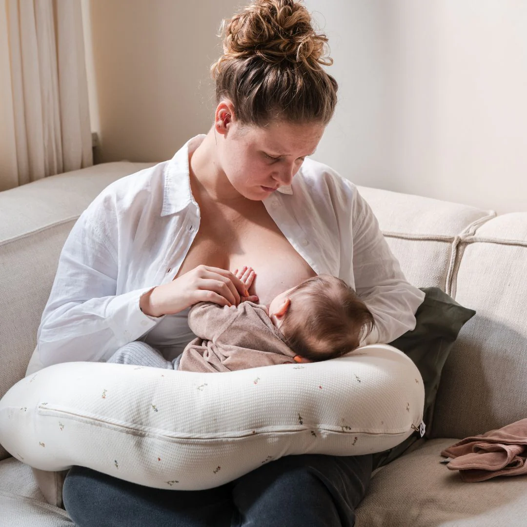Mãe a amamentar bebé usando almofada de amamentação branca com padrões pequenos em sofá cinza