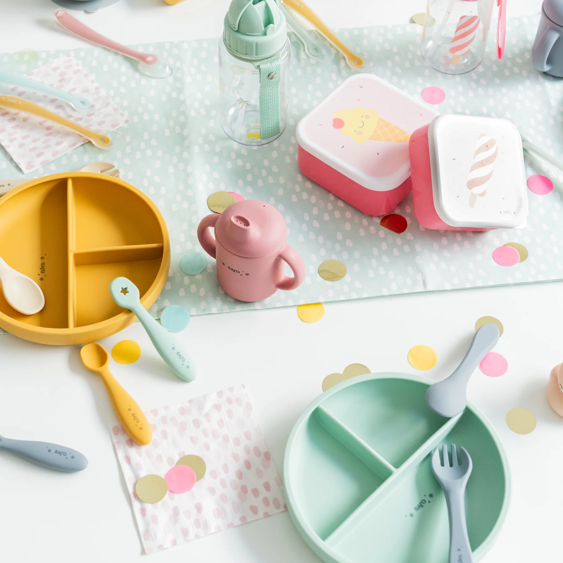 Utensílios de alimentação para bebés coloridos dispostos em mesa decorada com confetes, incluindo pratos divididos, talheres, copo e caixas de plástico