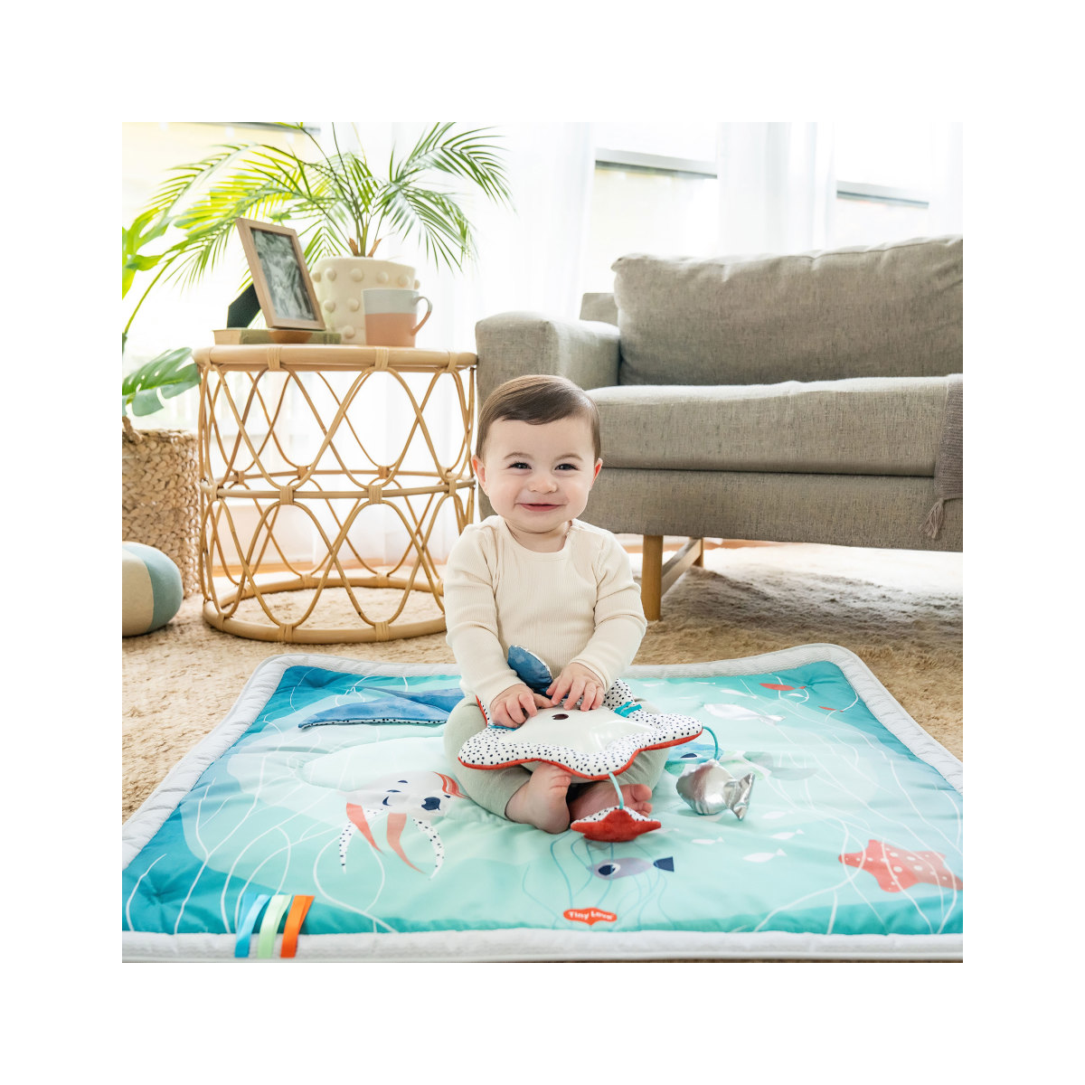 Bebé sentado em tapete azul com desenhos de animais marinhos, a brincar numa sala aconchegante com sofá cinzento e mesa de bambu.