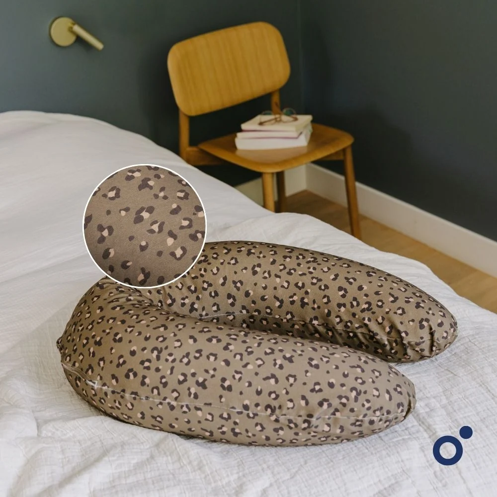 Almofada em forma de U com padrão de leopardo castanho em cama branca e cadeira de madeira no fundo.