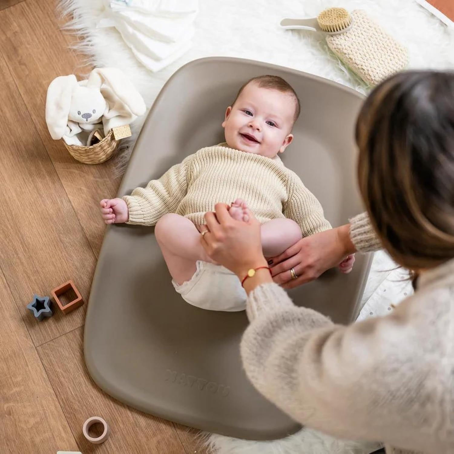 Bebé deitado num tapete cinza de mudar fraldas rodeado por brinquedos e uma pessoa a segurar os seus pés