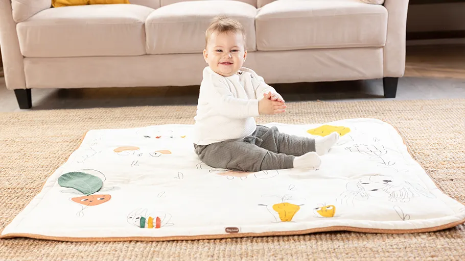 Bebé sentado numa manta colorida em contexto de sala com sofá e tapete
