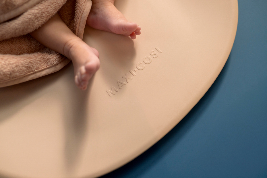 Bebé em manta castanha numa superfície redonda bege com marca MAXICOSI sobre fundo azul