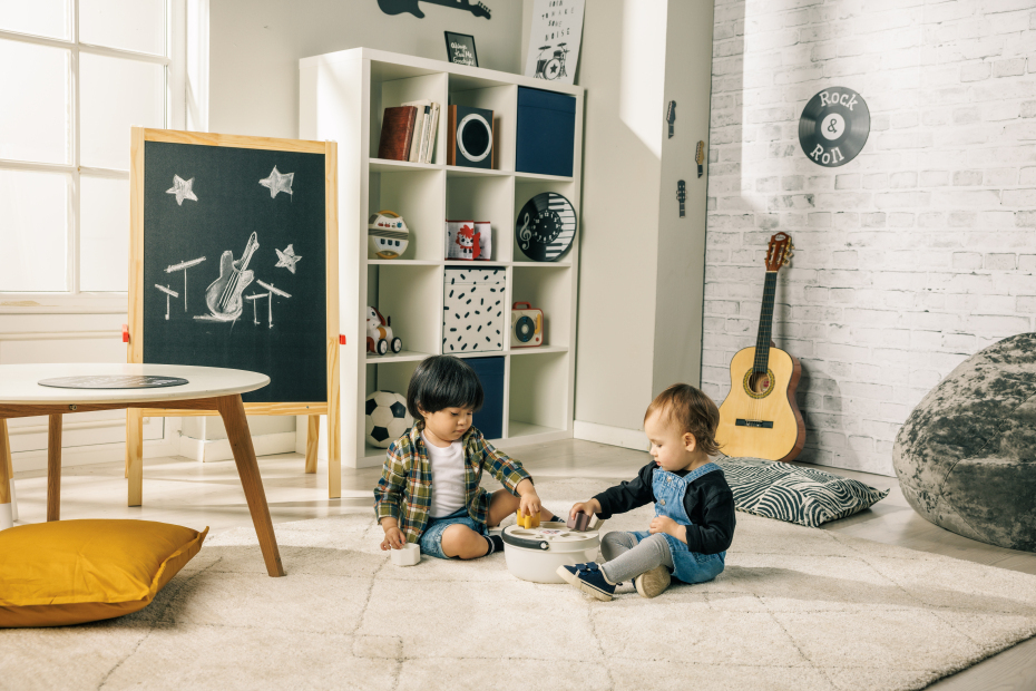 Dois bebés brincando numa sala infantil com tapete bege, almofadas, estante branca e guitarra encostada à parede.