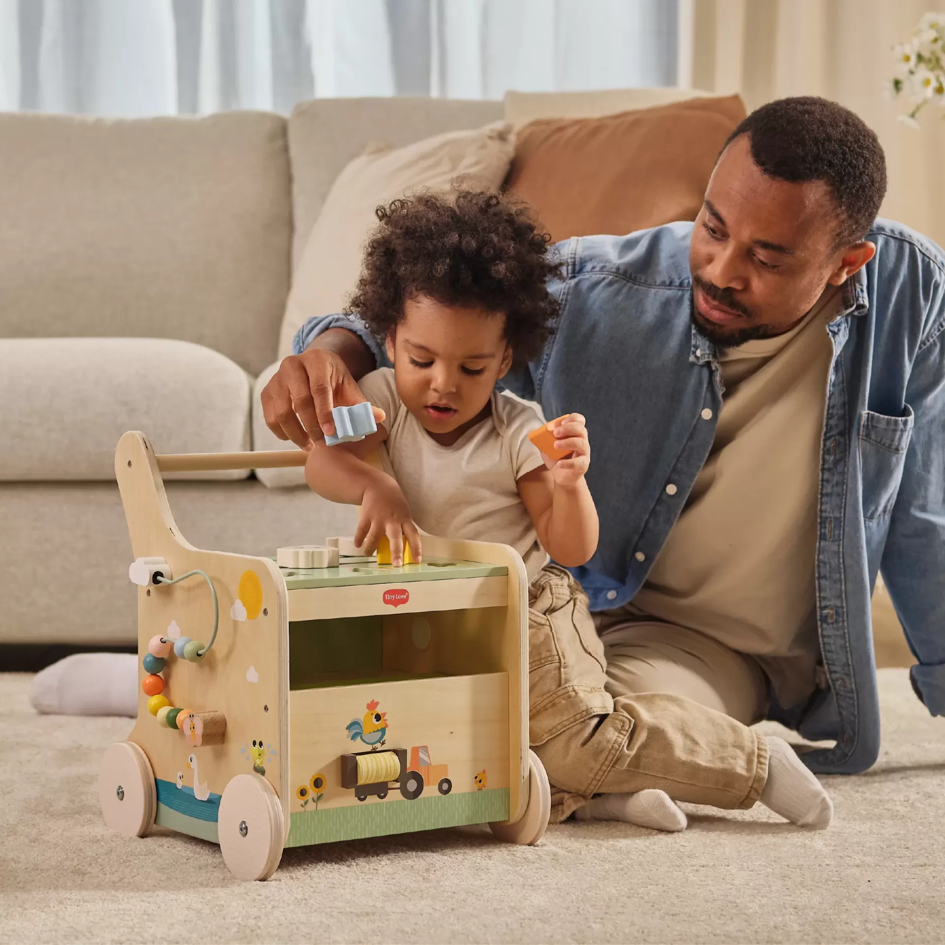Criança a brincar com carrinho de madeira colorido acompanhada por adulto em sala de estar