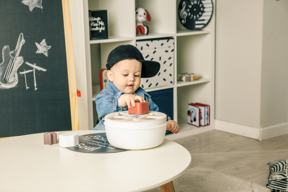 Criança pequena brinca com blocos de construção numa mesa redonda branca, com estante e quadro-negro ao fundo
