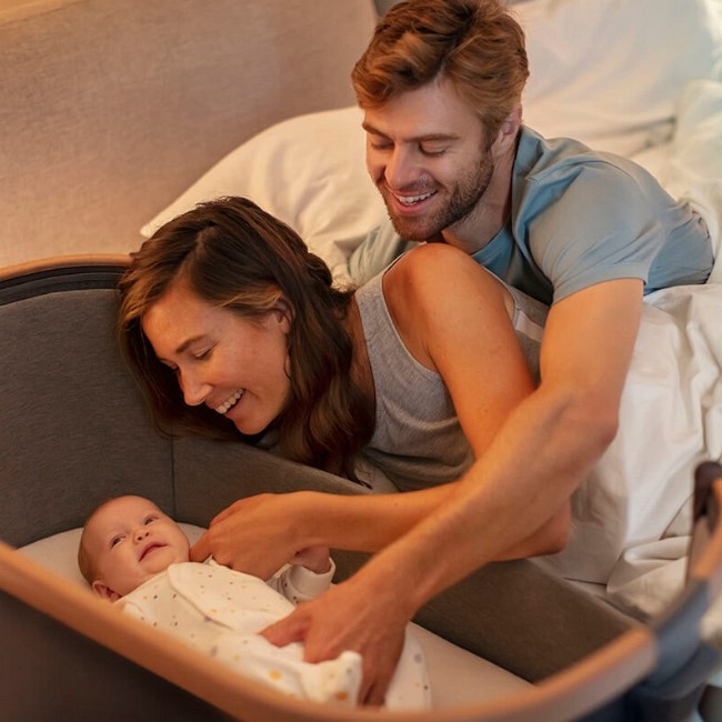 Berço cinza escuro com bebé e pais sorrindo numa cama