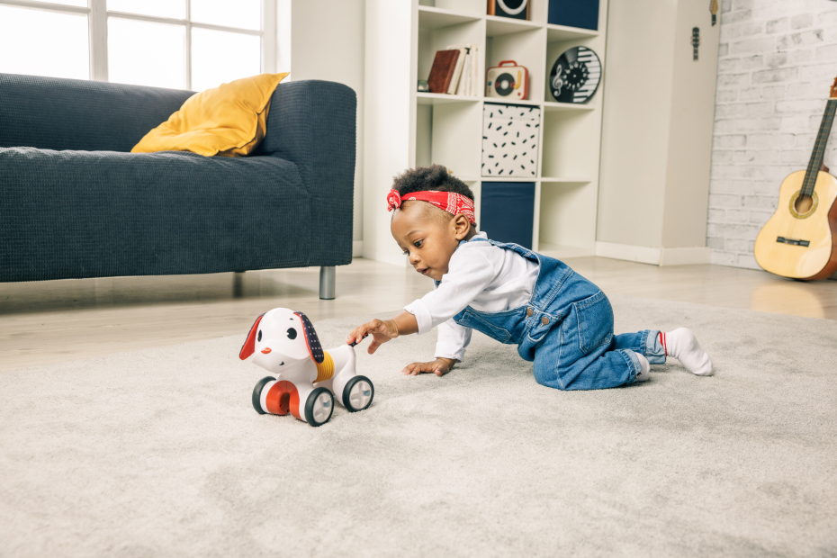 Criança a brincar com brinquedo de cão com rodas, chão cinza, sofá azul escuro e almofada amarela