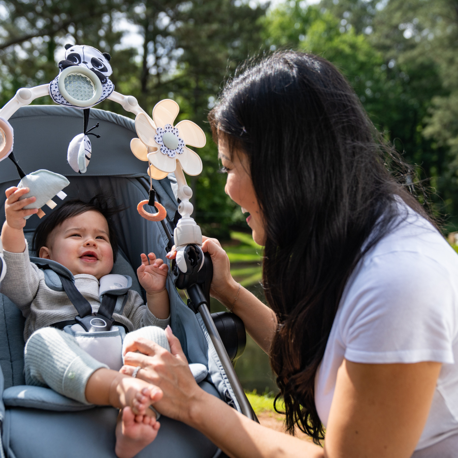 Bebé no carrinho cinzento com brinquedos e mulher interagindo ao ar livre