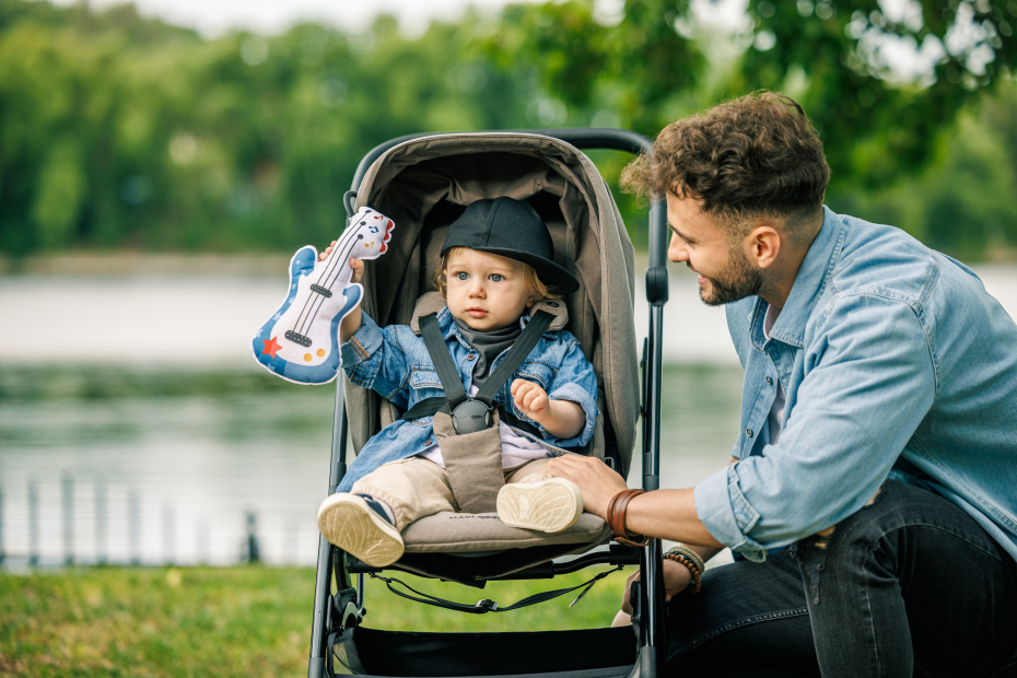 Bebé num carrinho cinza a segurar brinquedo guitarra, homem ao lado em parque