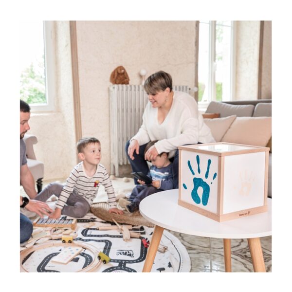 Cubo luminoso infantil de madeira com mãos coloridas numa mesa branca e família a brincar no tapete com carros de madeira.