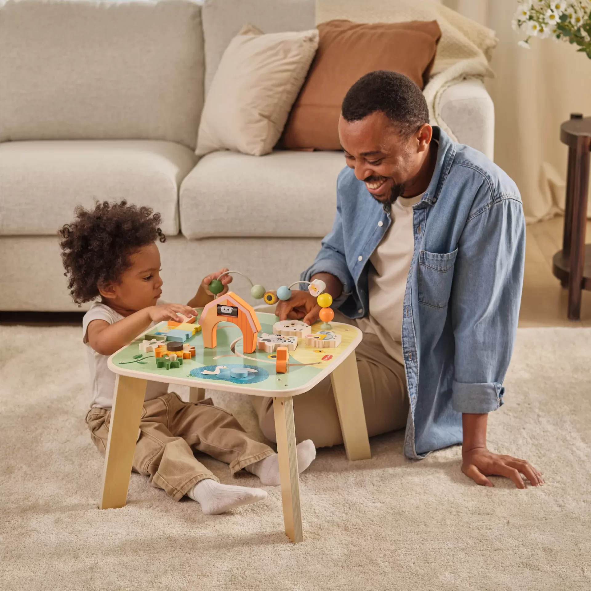Mesa de atividades infantil em madeira suave com peças coloridas, criança e adulto a brincar no tapete.