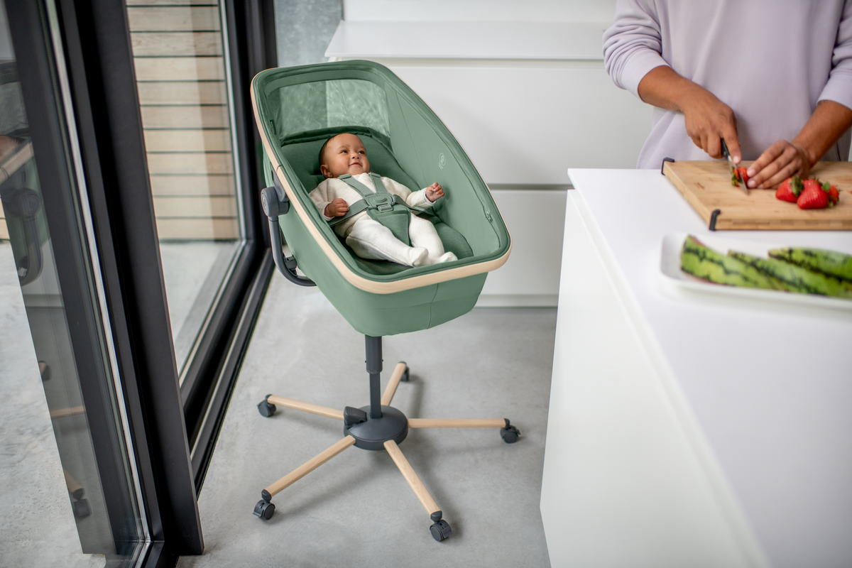 Berço portátil verde com base de madeira e rodas junto a janela e bancada branca