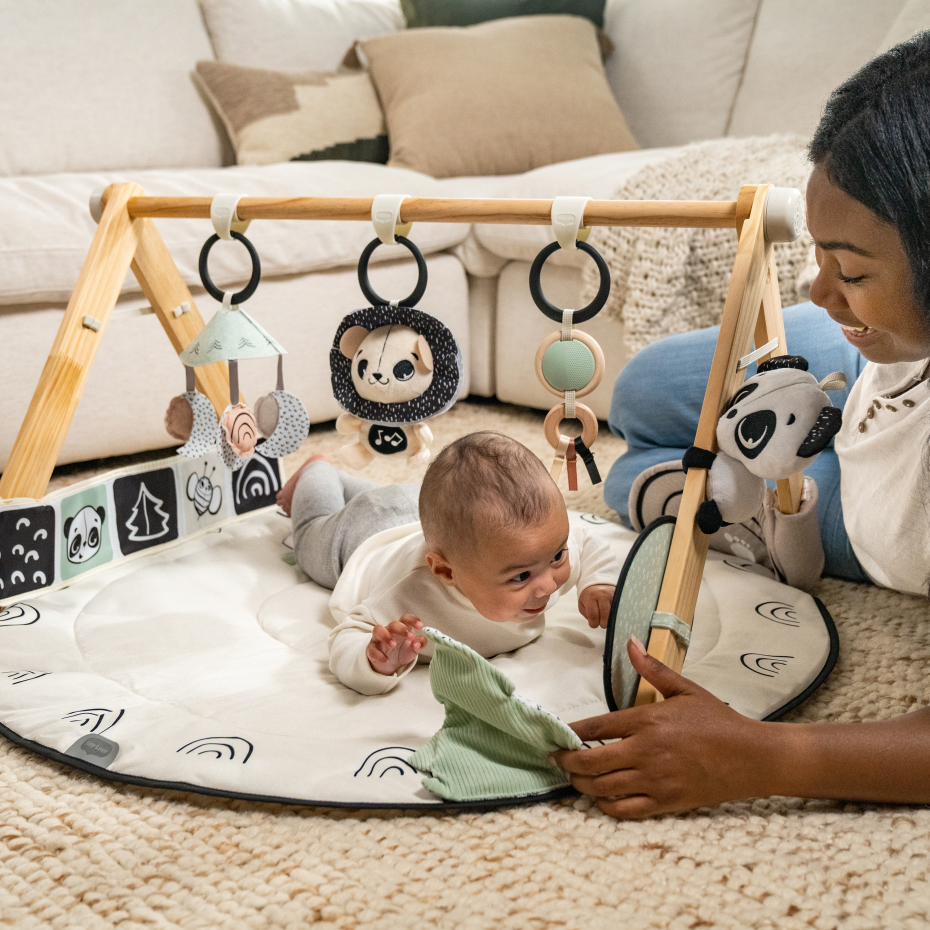 Tapete de atividades para bebé com brinquedos pendurados em estrutura de madeira num ambiente interior acolhedor.