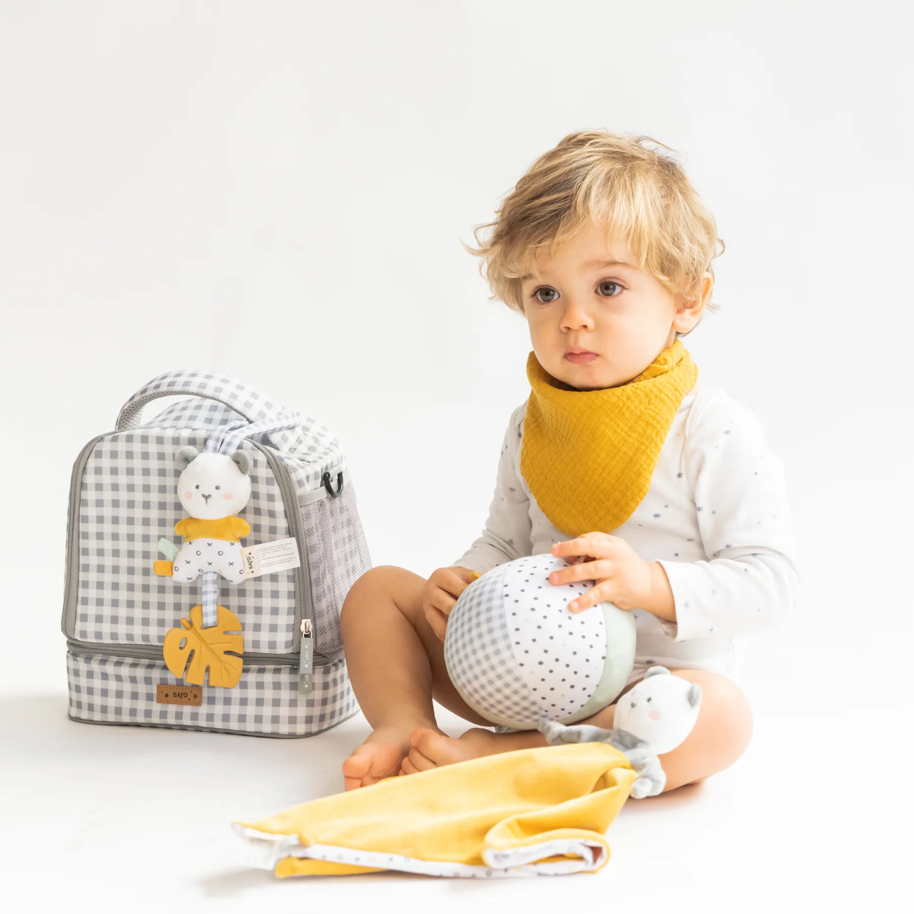Criança com babete amarelo, bola branca, mochila cinza xadrez e mantinha amarela