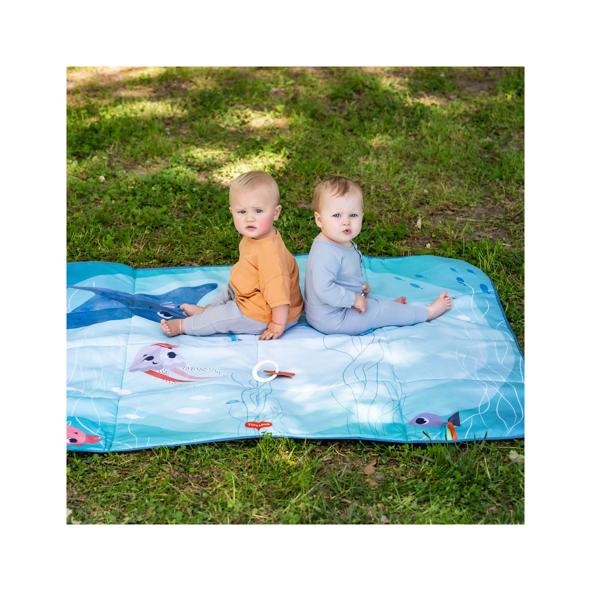 Bebés sentados num tapete de atividades azul claro com desenhos de peixes sobre relva ao ar livre