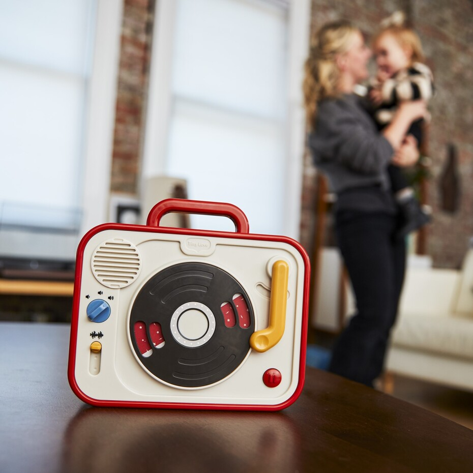 Leitor de música vintage em plástico vermelho e branco numa mesa com mulher a segurar criança desfocada ao fundo