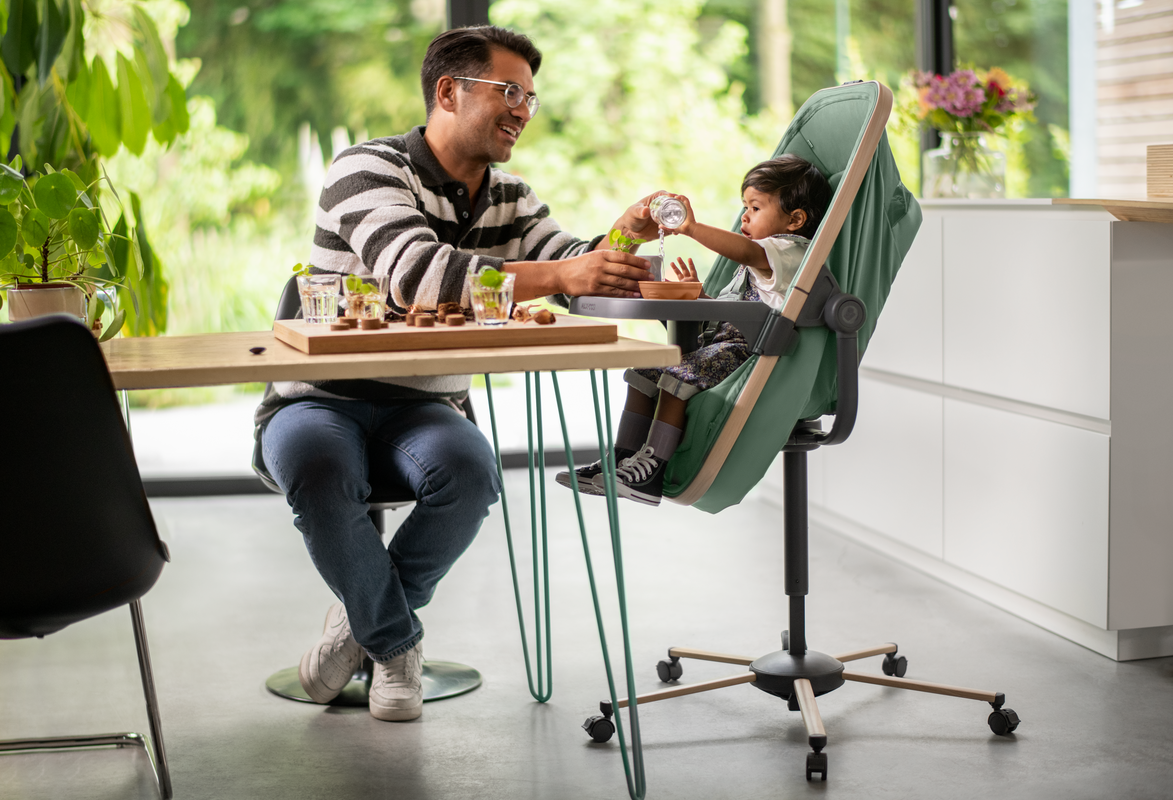 Cadeira alta infantil verde com criança e homem interagindo ao lado de mesa de madeira em ambiente moderno