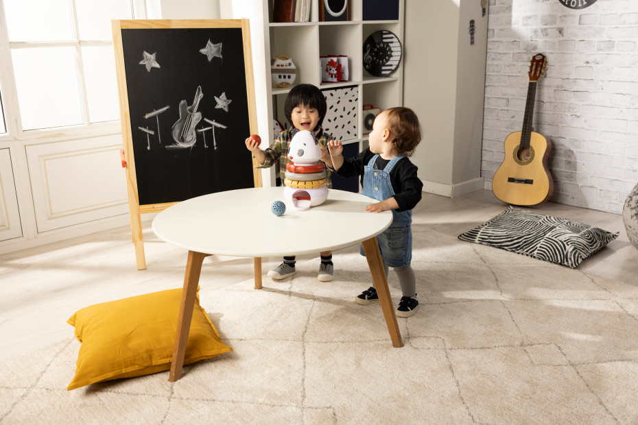 Sala de estar com tapete bege, mesa branca, quadro-negro desenhado, guitarra e duas crianças com brinquedo robô.