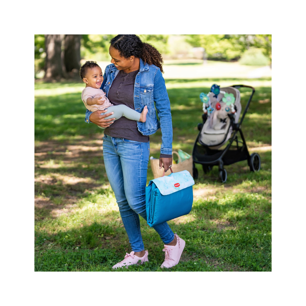 Mulher com mala térmica azul e branca segurando bebé, carrinho ao fundo em parque