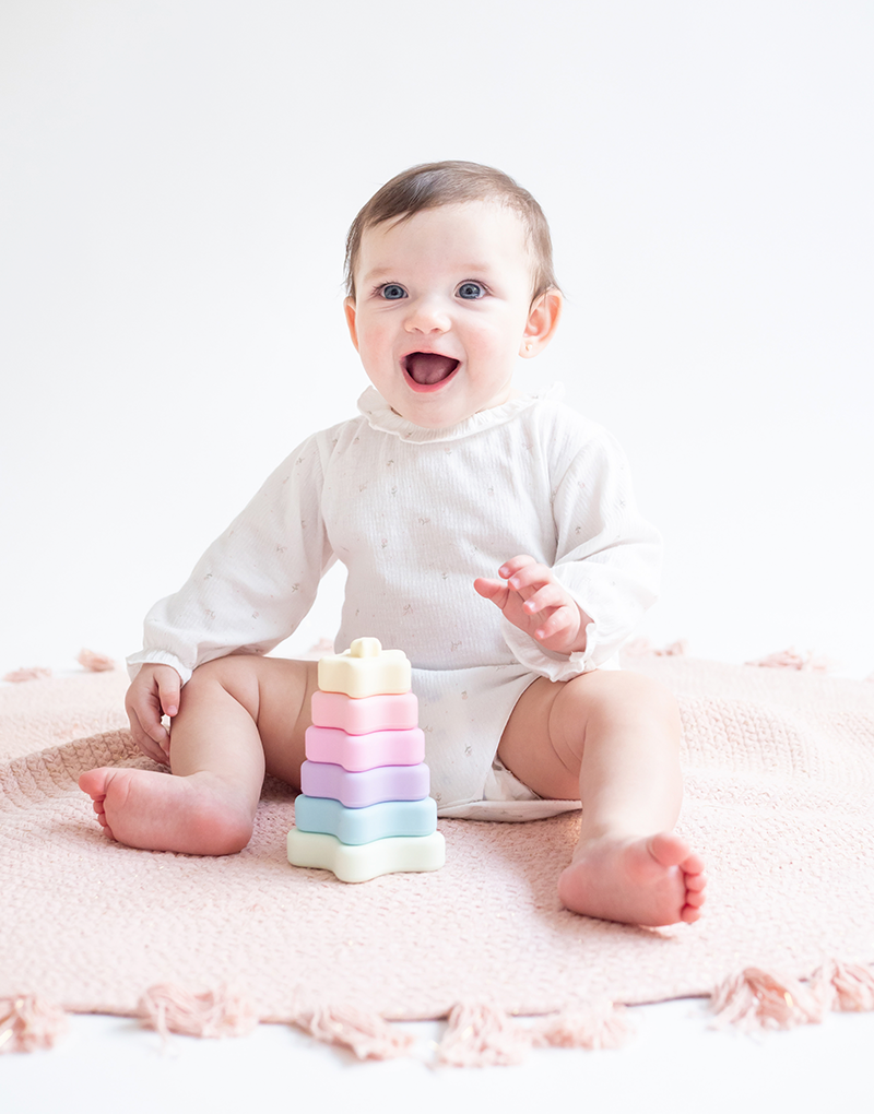 Bebé com body branco a brincar com brinquedo empilhável colorido em tapete cor-de-rosa claro.