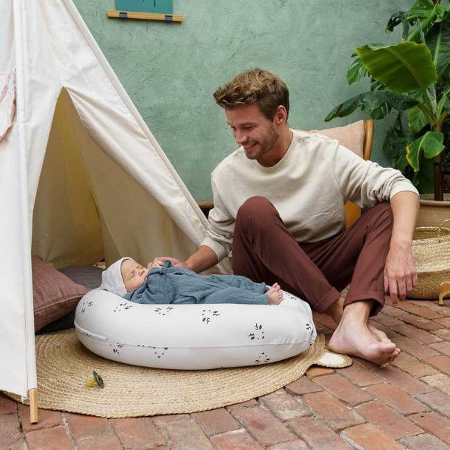 Almofada de amamentação branca com padrões pretos, bebé e homem sentados no chão de tijolos ao lado de tenda branca e plantas.