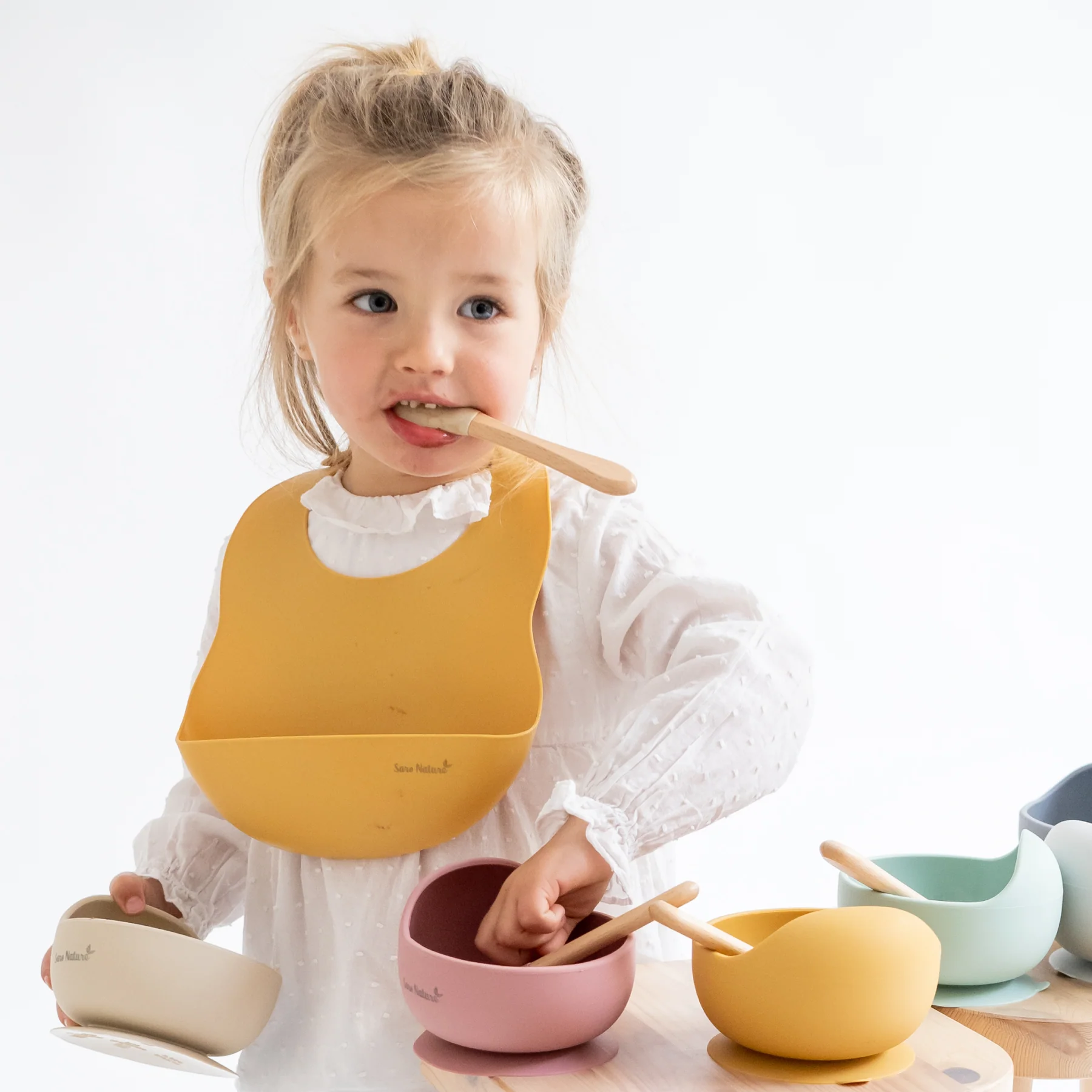 Criança com babete amarelo e tigelas coloridas com colheres.