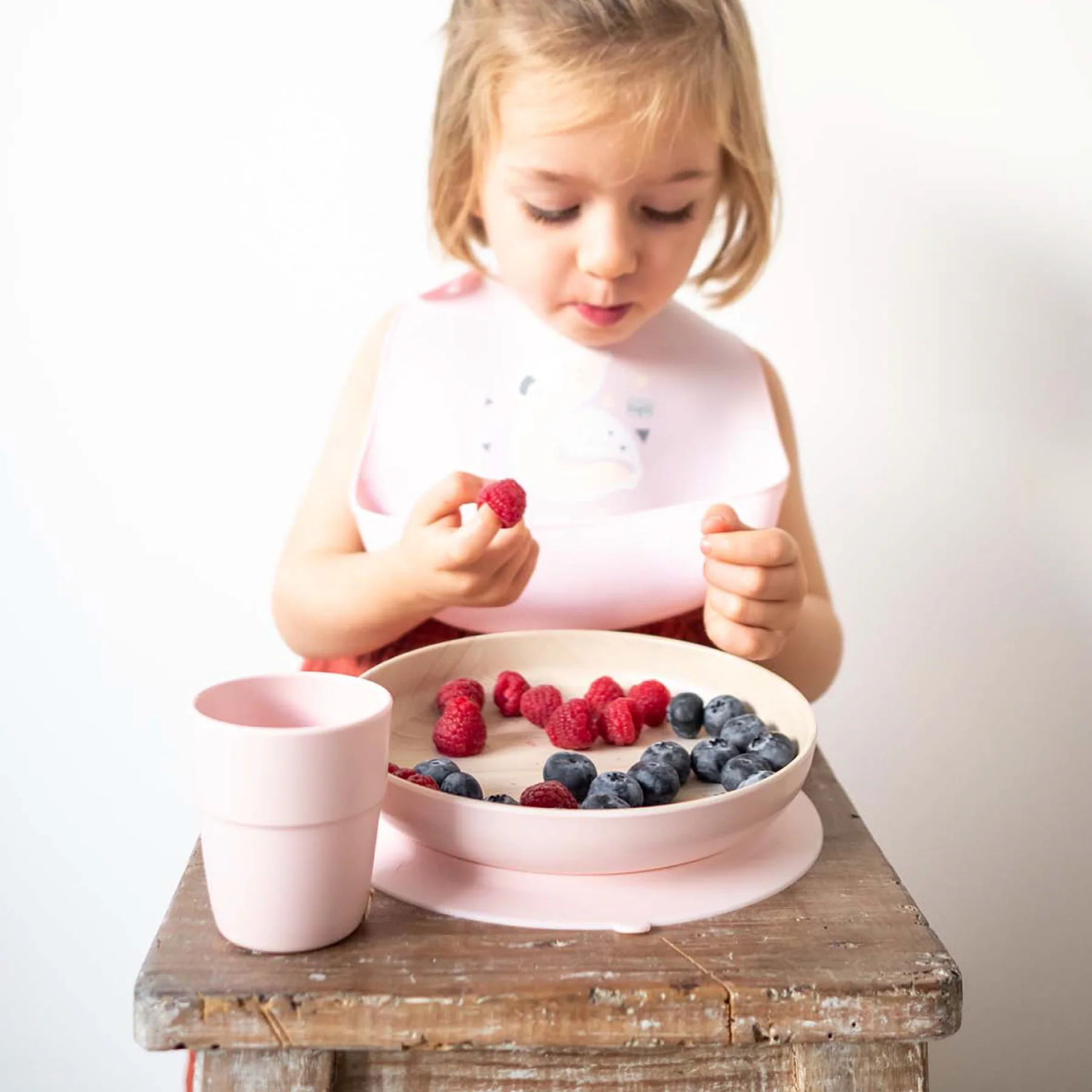 Menina sentada com babete rosa e prato rosa contendo frutas vermelhas e azuladas sobre mesa de madeira