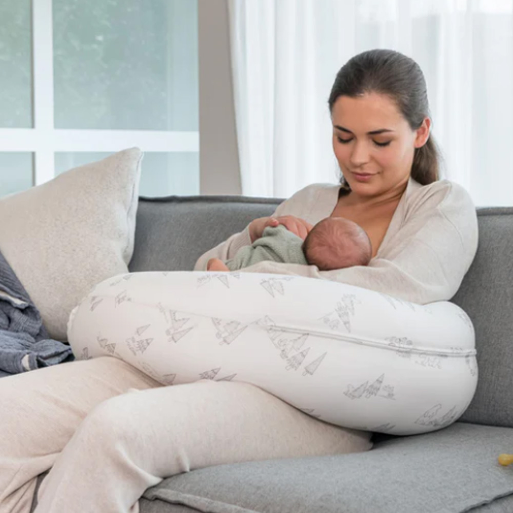 Almofada de amamentação branca com padrão de tenda cinza claro usada para segurar bebé bebido por mulher sentada num sofá cinza.