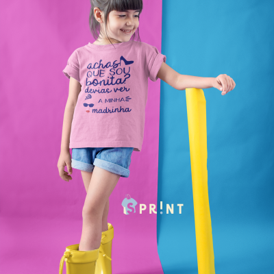 Camisola rosa para criança com texto em português e botas amarelas sobre fundo roxo e azul.