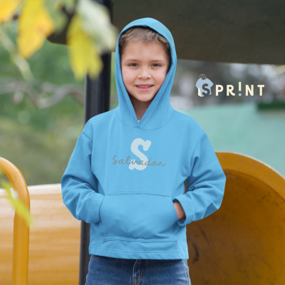 Menino com hoodie azul claro com 'S' e 'Salvador' em dourado, ao ar livre, com objetos amarelos ao fundo