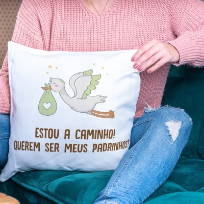 Travesseiro branco com estampa de cegonha e texto em castanho, segurado por pessoa sentada em sofá verde-azul.