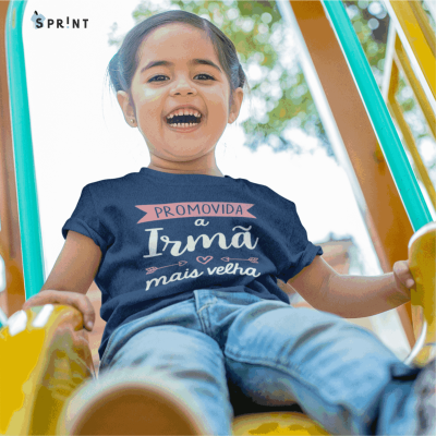 Criança sorridente no escorrega com camisola azul escura escrita 'PROMOVIDA a Irmã mais velha'.