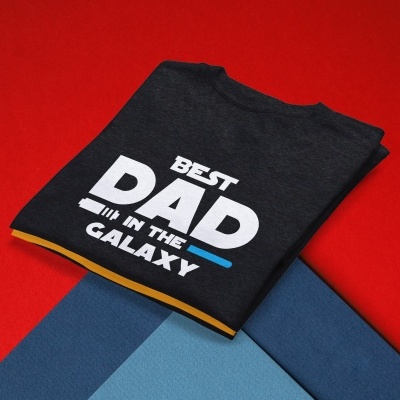 Camisola preta com texto branco 'BEST DAD IN THE GALAXY' sobre superfícies coloridas vermelho e azul.