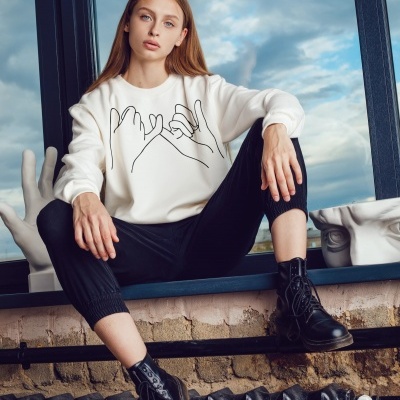 Moça loira em peitoril de janela com sweat branco, calças pretas e botas pretas